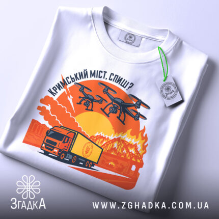 Футболка кримський міст горить, біла, складена на поверхні, фон нейтральний, принт зображає вантажівку та дрони на фоні палаючого мосту з оранжевими та червоними відтінками, з етикеткою на рукаві.