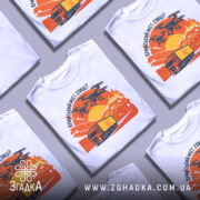 Футболка кримський міст горить, біла, зображена на манекені, фон нейтральний, принт зображає вантажівку та дрони на фоні палаючого мосту з оранжевими та червоними відтінками.