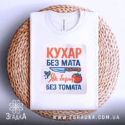 Футболка кухар без мата, біла, зображена зверху на плетеній основі, фон світло-сірий, принт містить текст "КУХАР БЕЗ МАТА" з ілюстрацією ножа та помідора, футболка складена.