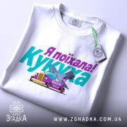 Футболка кукуха поїхала, біла, зображена з верхнього ракурсу, на фоні світло-сірого кольору, принт містить текст "Я поїхала! Кукуха" та малюнок автомобіля з птахом, на комірці є етикетка.