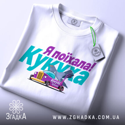 Футболка кукуха поїхала, біла, зображена з верхнього ракурсу, на фоні світло-сірого кольору, принт містить текст "Я поїхала! Кукуха" та малюнок автомобіля з птахом, на комірці є етикетка.