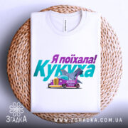 Футболка кукуха поїхала, біла, зображена зверху на плетеному кошику, на фоні світло-сірого кольору, принт містить текст "Я поїхала! Кукуха" та малюнок автомобіля з птахом.