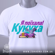 Футболка кукуха поїхала, біла, зображена з верхнього ракурсу, на фоні світло-сірого кольору, принт містить текст "Я поїхала! Кукуха" та малюнок автомобіля з птахом, на комірці є етикетка.