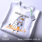 Футболка кукуха сказала поїхали. Біла футболка, складена на поверхні, з принтом птаха в халаті, що тримає планшет. На футболці видно текст 'Поїхали!'. Фон світлий, з легким відблиском, на футболці є етикетка.