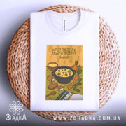 Футболка куліш білого кольору, зображена зверху, на фоні плетеного кошика. Принт з ілюстрацією кулішу, що містить каструлю з їжею, овочі та хліб, оформлений у яскравих кольорах.