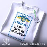 Футболка кум молодець. Біла футболка, зображена зверху, на фоні світло-сірого градієнту. Принт з текстом 'ГРАМОТА КУМ, ІБАТЬ ТИ МОЛОДЕЦЬ!' у синьому та чорному кольорах, герб України та жовто-блакитні стрічки. На футболці видно етикетку з зеленим шнурком.