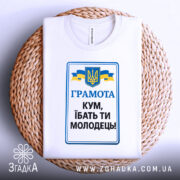 Футболка кум молодець. Біла футболка, зображена зверху, на фоні плетеного кошика. Принт містить текст 'ГРАМОТА КУМ, ІБАТЬ ТИ МОЛОДЕЦЬ!' у синьому та чорному кольорах, герб України та жовто-блакитні стрічки.