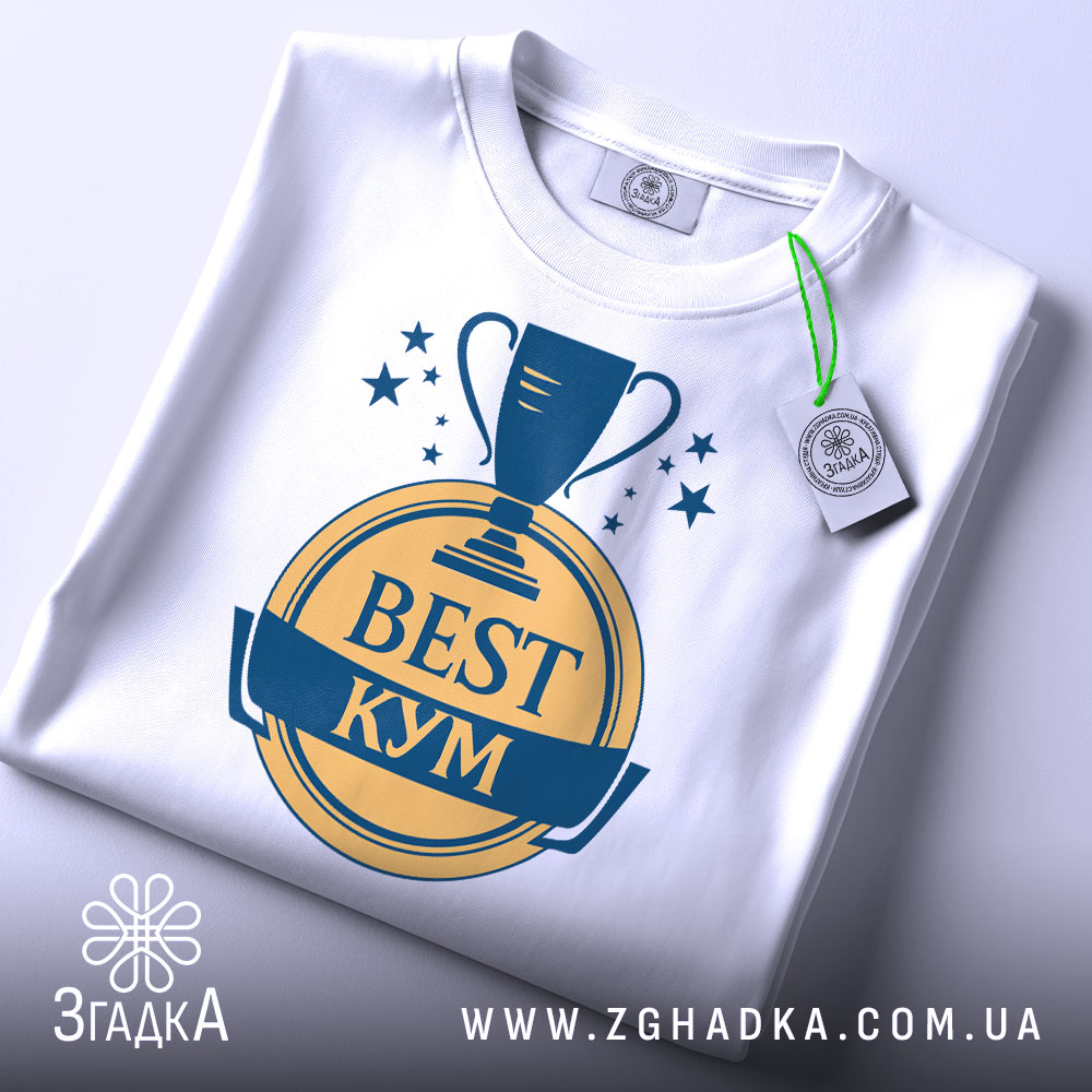 Футболка кум найкращий, біла, зображена зверху, складена на поверхні, фон світло-сірий, принт з трофеєм та написом 'BEST КУМ' у жовтому та синьому кольорах, з етикеткою зеленого кольору.