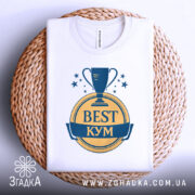 Футболка кум найкращий, біла, зображена зверху, складена на плетеній кошику, фон білий, принт з трофеєм та написом 'BEST КУМ' у жовтому та синьому кольорах.