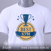 Футболка кум найкращий, біла, зображена зверху, складена на поверхні, фон світло-сірий, принт з трофеєм та написом 'BEST КУМ' у жовтому та синьому кольорах, з етикеткою зеленого кольору.