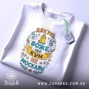 Футболка кум не москаль, біла, складена на поверхні, фон світло-сірий, принт з текстом "Дякую, Боже, що кум не москаль!" в яскравих кольорах, з етикеткою на зеленій стрічці.