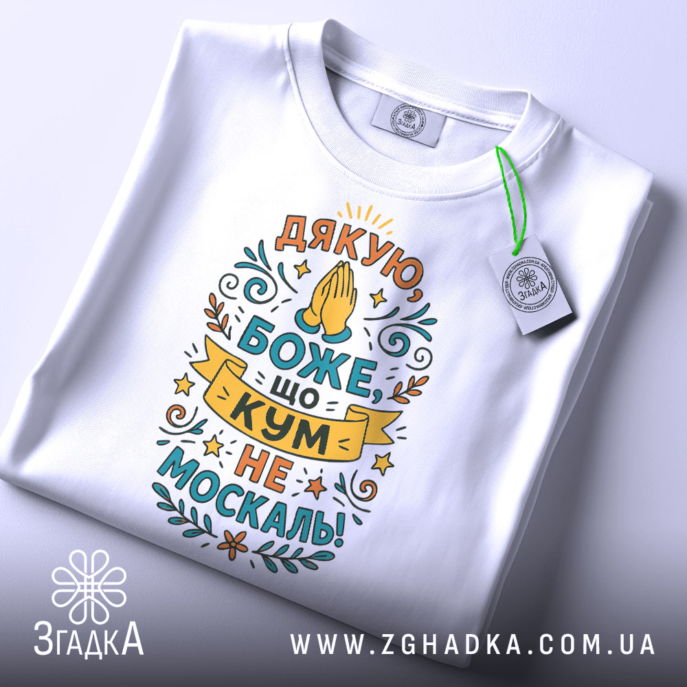 Футболка кум не москаль, біла, складена на поверхні, фон світло-сірий, принт з текстом "Дякую, Боже, що кум не москаль!" в яскравих кольорах, з етикеткою на зеленій стрічці.
