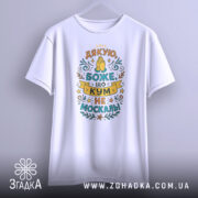 Футболка кум не москаль, біла, підвішена на вішалці, фон світло-сірий, принт з текстом "Дякую, Боже, що кум не москаль!" в яскравих кольорах, з елементами декору.