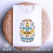 Футболка кум не москаль, біла, складена на плетеній кошику, фон білий, принт з текстом "Дякую, Боже, що кум не москаль!" в яскравих кольорах.