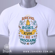 Футболка кум не москаль, біла, складена на поверхні, фон світло-сірий, принт з текстом "Дякую, Боже, що кум не москаль!" в яскравих кольорах, з етикеткою на зеленій стрічці.