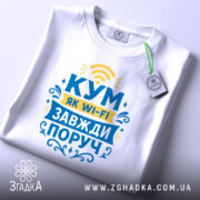 Футболка кум завжди поруч, біла, зображена зверху, фон світлий, принт з текстом "КУМ ЯК WI-FI ЗАВЖДИ ПОРУЧ" у жовтому та блакитному кольорах, на футболці є етикетка з логотипом.