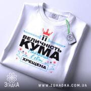 Футболка кума хрещена білого кольору, складена на поверхні, фон світло-сірий, принт з текстом "ВЕЛИЧНІСТЬ КУМА Найкраща в світі ХРЕЩЕНА" з червоною короною, зелена етикетка прикріплена до коміра.