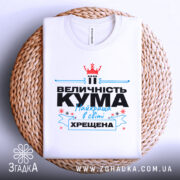 Футболка кума хрещена білого кольору, складена на плетеній кошику, фон світло-сірий, принт містить текст "ВЕЛИЧНІСТЬ КУМА Найкраща в світі ХРЕЩЕНА" з червоною короною та блакитними елементами.