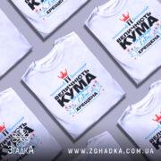 Футболка кума хрещена білого кольору, зображена з фронтального ракурсу на манекені, фон світло-сірий, принт містить текст "ВЕЛИЧНІСТЬ КУМА Найкраща в світі ХРЕЩЕНА" з червоною короною та блакитними елементами.