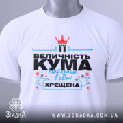 Футболка кума хрещена білого кольору, складена на поверхні, фон світло-сірий, принт з текстом "ВЕЛИЧНІСТЬ КУМА Найкраща в світі ХРЕЩЕНА" з червоною короною, зелена етикетка прикріплена до коміра.