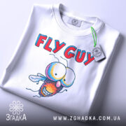 Футболка кумедна муха білого кольору, складена на столі, фон світло-сірий, принт з мультяшною мухою та текстом 'FLY GUY', на футболці є етикетка з зеленим шнурком.