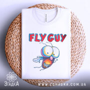 Футболка кумедна муха білого кольору, складена на плетеній кошику, фон світло-сірий, принт з мультяшною мухою та текстом 'FLY GUY', видима етикетка на комірці.