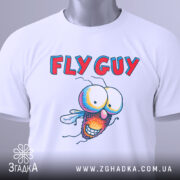 Футболка кумедна муха білого кольору, складена на столі, фон світло-сірий, принт з мультяшною мухою та текстом 'FLY GUY', на футболці є етикетка з зеленим шнурком.