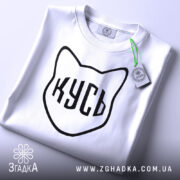 Футболка кусь білого кольору, складена на столі, з видимим ярликом, фон світлий, принт у вигляді котячої голови з написом 'КУСЬ' чорного кольору.