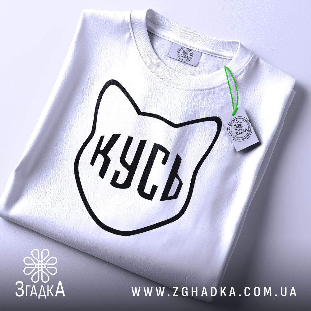 Футболка кусь білого кольору, складена на столі, з видимим ярликом, фон світлий, принт у вигляді котячої голови з написом 'КУСЬ' чорного кольору.