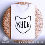 Футболка кусь білого кольору, складена на кошику з ротанга, фон світлий, принт у вигляді котячої голови з написом 'КУСЬ' чорного кольору.