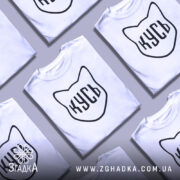 Футболка кусь білого кольору, зображена з фронтального ракурсу на манекені, фон світлий, принт у вигляді котячої голови з написом 'КУСЬ' чорного кольору.