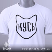 Футболка кусь білого кольору, складена на столі, з видимим ярликом, фон світлий, принт у вигляді котячої голови з написом 'КУСЬ' чорного кольору.