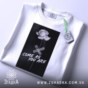 Футболка квітка монохром, біла, складена на поверхні, фон світло-сірий, принт з чорним фоном, білою трояндою та написом 'COME AS YOU ARE', з етикеткою зеленого кольору.