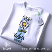 Футболка квітка ромашка, біла, складена на поверхні, злегка з верхнього ракурсу, фон світлий, принт з ромашкою та написом 'GOOD VIBES' з елементами кольору.