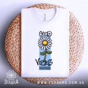 Футболка квітка ромашка, біла, складена на плетеній кошику, фон світлий, принт з ромашкою та написом 'GOOD VIBES' з елементами кольору.