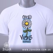 Футболка квітка ромашка, біла, складена на поверхні, злегка з верхнього ракурсу, фон світлий, принт з ромашкою та написом 'GOOD VIBES' з елементами кольору.