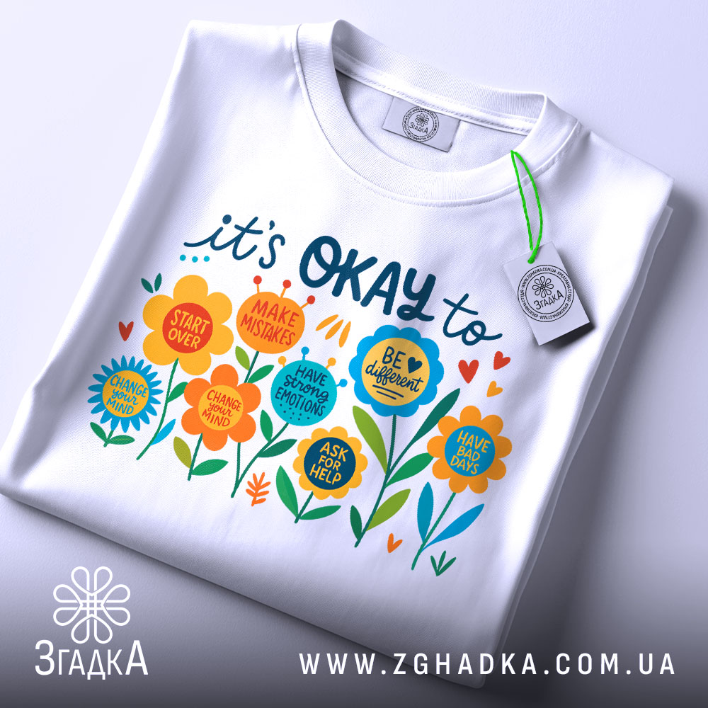 Футболка квіткова мотивація. Біла футболка, складена на столі, з кольоровим принтом, видимим спереду. Принт містить яскраві квіти та текст, що підтримує позитивні емоції. На футболці є етикетка з зеленим шнурком. Фон світло-сірий, що підкреслює деталі принта.