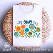 Футболка квіткова мотивація. Біла футболка, складена на плетеній кошику, з кольоровим принтом, що містить квіти та текст. Принт яскравий, з позитивними повідомленнями. Фон світло-сірий, що підкреслює деталі принта.