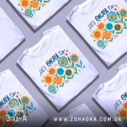 Футболка квіткова мотивація. Біла футболка з кольоровим принтом, зображеним на грудях, на манекені з металевим блиском. Принт містить різнокольорові квіти та текст, що закликає до позитивного мислення. Фон світло-сірий, що підкреслює яскравість кольорів принта.