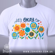 Футболка квіткова мотивація. Біла футболка, складена на столі, з кольоровим принтом, видимим спереду. Принт містить яскраві квіти та текст, що підтримує позитивні емоції. На футболці є етикетка з зеленим шнурком. Фон світло-сірий, що підкреслює деталі принта.