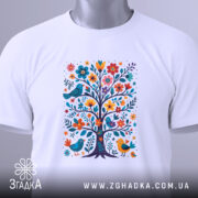 Футболка квіткове дерево, біла, складена на столі, фон світлий, принт з кольоровим деревом, птахами та квітами, з етикеткою на зеленому шнурку.