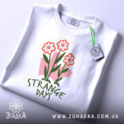 Футболка квіткові арт дні, біла, зображена зверху, складена на поверхні, фон світло-сірий, принт з трьома червоними квітами та написом 'STRANGE DAYS' зеленого кольору, з етикеткою.