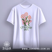 Футболка квіткові арт дні, біла, підвішена на вішалці, фон світло-сірий, принт з трьома червоними квітами та написом 'STRANGE DAYS' зеленого кольору.