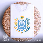 Футболка квітковий тризуб, біла, складена на кошику з плетеного матеріалу, фон світло-сірий, принт у вигляді тризуба з квітковими елементами в жовтих, блакитних та зелених відтінках.