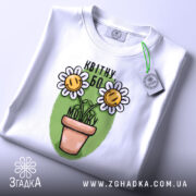 Футболка квітну бо можу, білого кольору, складена на столі, фон білий, принт з двома усміхненими соняшниками в горщику на зеленому фоні, з етикеткою зеленого кольору.