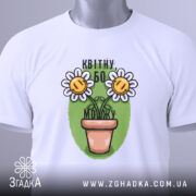 Футболка квітну бо можу, білого кольору, складена на столі, фон білий, принт з двома усміхненими соняшниками в горщику на зеленому фоні, з етикеткою зеленого кольору.