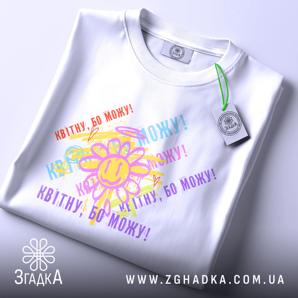 Футболка квітну бо можу, біла, зображена на плоскій поверхні, фон світло-сірий, принт з усміхненим сонечком та кольоровими написами, що повторюють фразу 'Квітну, бо можу!', з етикеткою на рукаві.