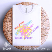 Футболка квітну бо можу, біла, зображена на плоскій поверхні, фон світло-сірий, принт з усміхненим сонечком та кольоровими написами, що повторюють фразу 'Квітну, бо можу!', на плетеній основі.