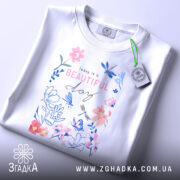 Футболка квіти прекрасний день, складена футболка на білому фоні, вид зверху, принт з квітами та метеликами в пастельних кольорах, текст "Today is a BEAUTIFUL day" розташований в центрі, з етикеткою на зеленому шнурку.