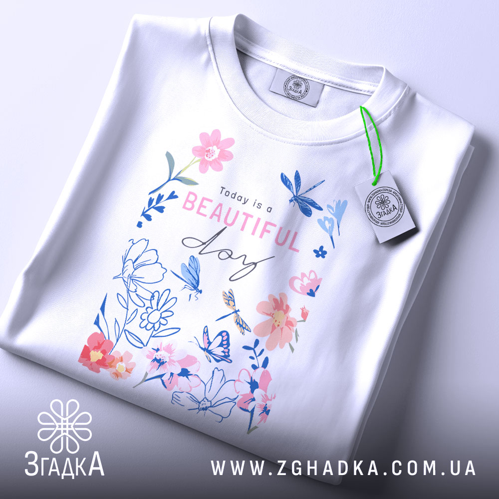 Футболка квіти прекрасний день, складена футболка на білому фоні, вид зверху, принт з квітами та метеликами в пастельних кольорах, текст "Today is a BEAUTIFUL day" розташований в центрі, з етикеткою на зеленому шнурку.